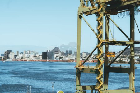 Foto de A shot of a yellow harbor crane in front of the cyan seawater, with cables, and staircases following on height of the equipment, and in the background the cityscape; a copy space area on the left - Imagen libre de derechos