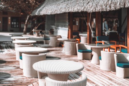 Téléchargez les photos : View of an empty outdoor terrace with cozy creamy-colored plastic wicker furniture seats with cushions, wooden flooring, and a rustic thatched roof create a charming atmosphere, perfect for unwinding - en image libre de droit