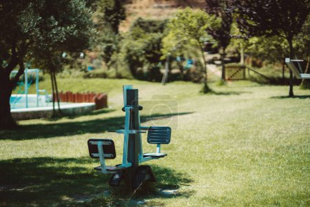 Téléchargez les photos : Close-up shot of an outdoor fitness machine in the focused foreground on a public park with children's playground equipment and trees spread throughout the park - en image libre de droit