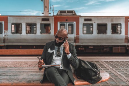 Photo for A black man multitasks on a blue-sky day, sitting on a wooden park bench while talking on the phone and jotting down notes. His weekender sits by his side, as a train buzzes by in the background. - Royalty Free Image