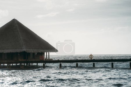 Photo for A close-up of an overwater bungalow, with a thatched roof, connected to a wooden walkway over water on a day with gray skies and calm seawater - Royalty Free Image