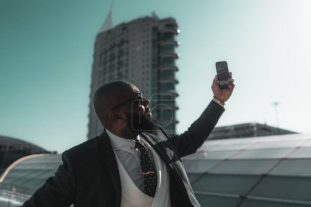 Téléchargez les photos : A portrait of a black, unshaven, and bald male, looking dapper in his tailored suit taking a selfie in front of an emblematic building of luxury housing, located in Lisbon, on a radiantly sunny day - en image libre de droit