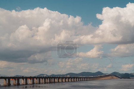 Téléchargez les photos : A view of the Rio Niteroi bridge spanning across the water contrasting against the vast white clouds above. In the background, majestic mountains can be seen, adding depth and texture to the landscape - en image libre de droit