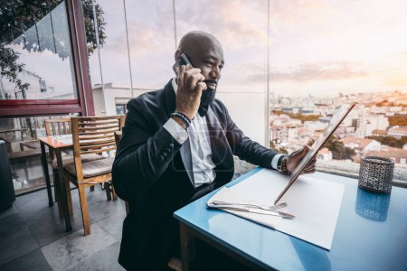 Téléchargez les photos : A stylish bald unshaven black male dressed in a sleek black suit is engaged in a phone conversation while perusing the menu at a restaurant located on a terrace overlooking a bustling cityscape - en image libre de droit