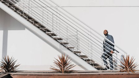 Téléchargez les photos : An African businessman starting to climb up the white staircase in front of the white wall, the man is wearing a grey suit in a minimalist aesthetic set with cactus decorating the scene - en image libre de droit