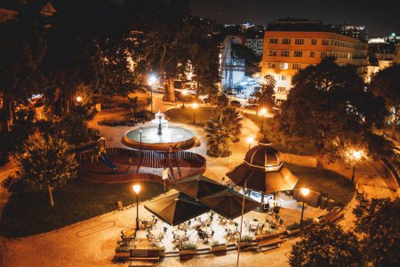 Téléchargez les photos : A top view of a well-lit urban garden at night. In the center, a fountain, a small playground with a visible slide, a terrace with tables and sunshades, and many trees surround the site - en image libre de droit