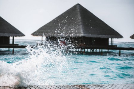 Téléchargez les photos : The shot is taken with a selective focus on the water splashing against a wooden footpath, leading to an overwater bungalows with an elegant design featuring thatched roofs, in the Maldives - en image libre de droit