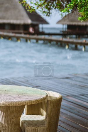 Photo for A vertical shot, with a cream-colored balcony table and chair in a rattan-style design and a tree branch with leaves taking center stage. The sea, the bungalows, and the boardwalk set the backdrop - Royalty Free Image