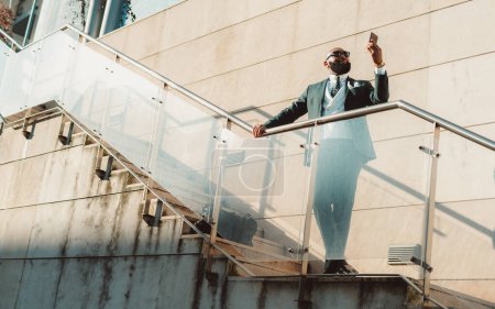 Photo for A well-dressed bald bearded black man wearing a suit and sunglasses, looking at his cell phone while standing on the stairway with a seamless glass railing with stone damp patches on a sunny day - Royalty Free Image