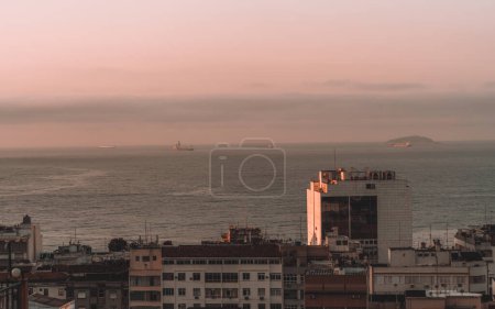 Foto de Foto de bajo perfil del océano mientras se mezcla con el cielo rosado en esta toma de la puesta del sol. Los edificios degradados en primer plano contrastan con el mar sereno, mientras que los barcos se desplazan pacíficamente a lo lejos. - Imagen libre de derechos