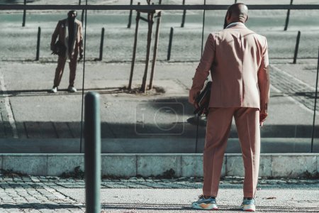 Téléchargez les photos : Un cliché d'un homme noir élégant au dos chauve dans un costume de couleur crème avec une mallette à la main et des baskets à la mode, attendant au passage supérieur a son image reflétée dans le bâtiment en face - en image libre de droit