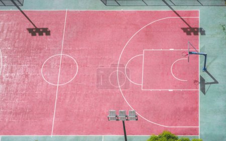 Foto de An aerial view of a red basketball court with some silhouettes of the LED vertical spotlights and the basket; view from high above of red and green basketball playground field on a sunny day - Imagen libre de derechos