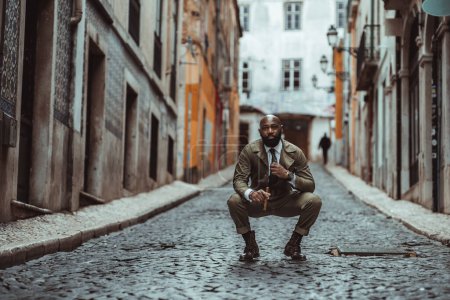 Photo for A shot focused on a bald black man dressed in khaki overalls standing in the middle of a black cobblestone street with typical old houses, crouching down, holding a cigar in shallow depth of field - Royalty Free Image