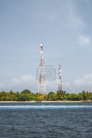 Foto de A vertical capture from a yacht overlooking the coast of a private island in the Maldives, on a blue sky day, overgrown with vegetation and two signal repeater towers. In the foreground little beach - Imagen libre de derechos