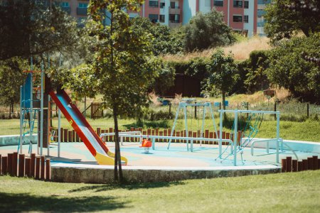 Photo for The capture of a children's playground, focusing on a colorful slide next to the swing and the climbing equipment in a round area partially enclosed by wood stakes - Royalty Free Image