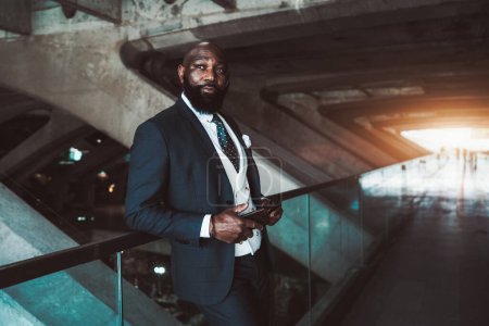 Téléchargez les photos : Un homme noir élégant vêtu d'un costume et d'une cravate à pois, appuyé avec désinvolture contre une balustrade en verre bleuté dans une station de transport en commun. Il tient son smartphone, suggérant qu'il attend peut-être - en image libre de droit