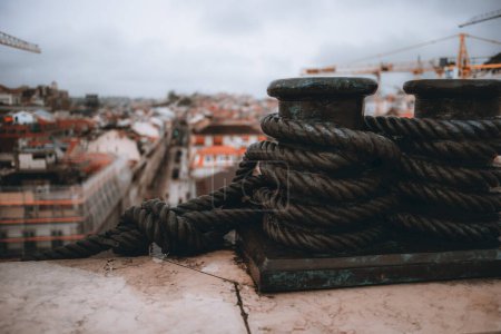 Téléchargez les photos : A close-up shot with a focus on a metal sculpture, in medium bronze color, of a bollard with a thick rope attached and around it, with the city of Lisbon out of focus in the background - en image libre de droit