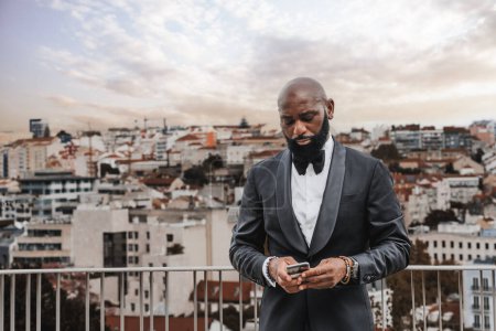 Téléchargez les photos : Portrait of a charming and gallant black man, well dressed in a black suit, a bow tie, and a white shirt, this one in a closed semblance looking at his smartphone on a terrace overlooking the city - en image libre de droit