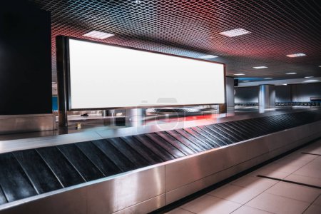 Photo for Shot in low-key of a very large rectangular blank billboard mockup facing a baggage claim in an airport terminal; a big white blank advertisement template indoors in front of a baggage handling system - Royalty Free Image
