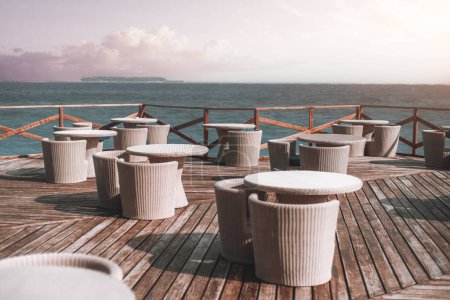 Photo for View of an serene outside cafe restaurant area filled by the beach ocean with rattan-effect tables and chairs on the wooden deck and fenced, in a peaceful background with lavender sky with clouds - Royalty Free Image