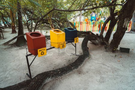 Téléchargez les photos : Three wooden recycling containers for waste collection are raised to human height on the white sand beach: the red for metal, yellow for plastic, and blue for paper, with a tree trunk right bellow - en image libre de droit