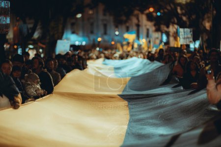 Foto de Lisboa, Portugal - 24 de febrero de 2023: En un disparo nocturno de bajo perfil, los manifestantes sostienen una gran bandera con un enfoque selectivo en su comienzo, marcando un aniversario de la guerra en el territorio de Ucrania. - Imagen libre de derechos