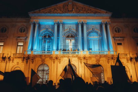 Photo for Lisbon, Portugal - February 24, 2023: The one-year anniversary of the war protest on a square, at night, as the building behind the crowed of protesters shines in the colors of the Ukrainian flag - Royalty Free Image