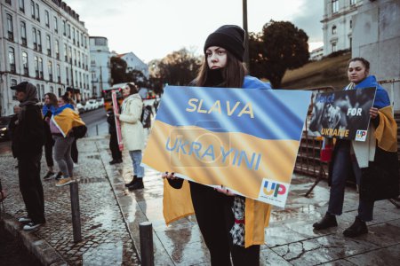 Foto de Lisboa, Portugal - 24 de febrero de 2023: En medio del frío, una manifestante se mantiene firme, agarrando un cartón de bandera ucraniana con un poderoso cartel contra la guerra, marcando un año desde que comenzó el conflicto.. - Imagen libre de derechos
