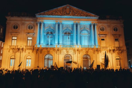 Foto de Lisboa, Portugal - 24 de febrero de 2023: Una foto nocturna del primer aniversario de la protesta de guerra en una plaza, mientras el edificio detrás de los manifestantes se ilumina con los colores de la bandera ucraniana - Imagen libre de derechos