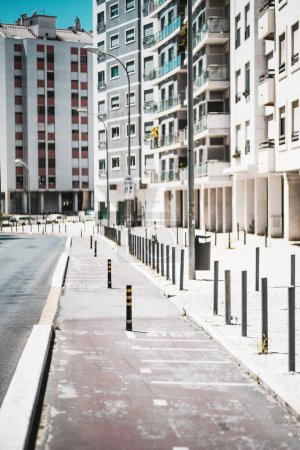 Photo for Vertical shot on a bright sunny day, a red degraded bicycle path runs between the road and the pedestrian walkway. Time and use have taken their toll on the path, next to residential buildings - Royalty Free Image