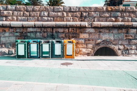 Foto de On the boardwalk of Cascais, a handful of eco-points are positioned against a sedimentary calcareous brownstone wall. They are located near an underground passageway with iron grates. - Imagen libre de derechos