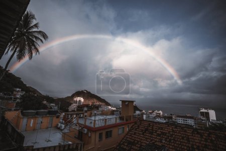 Téléchargez les photos : From a favela, a wide-angle low-key view of a balcony facing a backdrop of dark, moody skies; a beautiful rainbow shines through the clouds, adding a burst of color and hope to the scene; - en image libre de droit