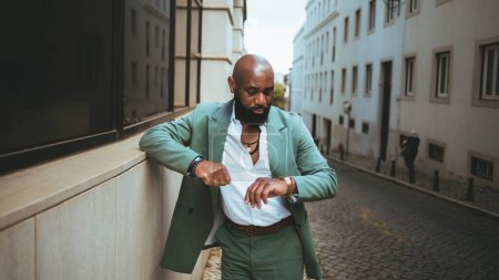 A sophisticated bald black man wearing a pastel green suit and a white shirt, accessorized with necklaces and checks the time on his wristwatch while standing in the middle of a cobblestone street