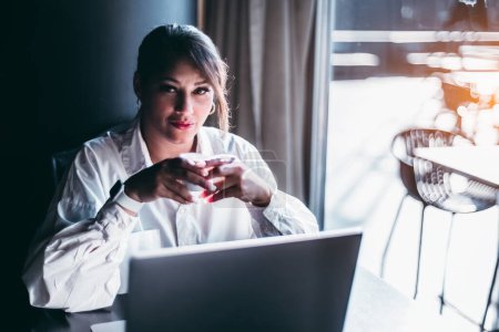 Téléchargez les photos : Une femme latina qui regarde séduisamment dans la caméra, elle est légèrement joufflue, tient une tasse de café pendant qu'elle prend une pause du travail, assise devant son ordinateur près d'une fenêtre ensoleillée portant un blanc - en image libre de droit
