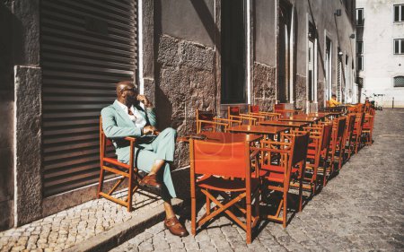Téléchargez les photos : Un élégant homme noir chauve est assis seul, perdu dans la pensée, sur une terrasse fermée à Lisbonne, Portugal ; le mobilier d'extérieur reste, brillant sous le soleil éclatant contre le trottoir traditionnel - en image libre de droit