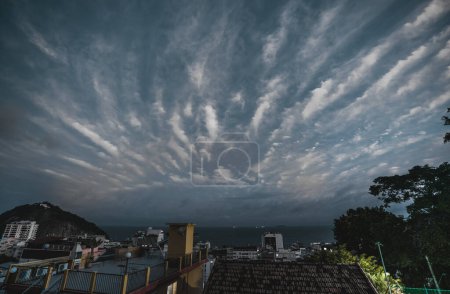 Téléchargez les photos : Depuis le toit d'une favela, une vue imprenable sur le quartier de Leme à Rio ; Le ciel bleu foncé est rempli de nuages qui créent des rayures fraîches, résultant en un paysage nuageux spectaculaire tourné dans un éclairage discret - en image libre de droit