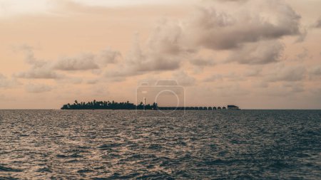 Foto de Disparo desde un barco navegando en el mar, esta puesta de sol de bajo perfil ofrece una vista fascinante de una isla en las Maldivas. Las siluetas de los bungalows en el fondo realzan la belleza de la escena - Imagen libre de derechos