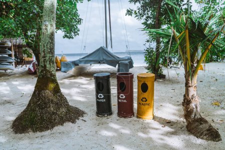 Téléchargez les photos : Trois bacs de recyclage cylindriques écologiques, colorés respectivement en jaune, rouge et bleu pour le plastique, le métal et le papier, ont été placés sur une plage entourée d'arbres tropicaux au feuillage varié. - en image libre de droit