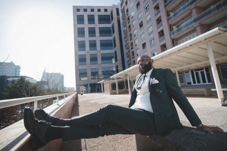 Photo for A well-dressed black man in a wide angle wears a full beard and relaxes; leaning his back, crossing his legs, and supporting himself with his arms as he enjoys the warmth of the sun on his face - Royalty Free Image