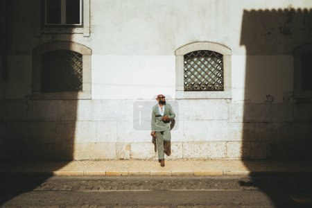 Téléchargez les photos : Un homme chauve noir semble confiant et prêt à se tenir debout dans son costume pastel vert contre le mur ; le séparer de la route est le trottoir, et les rayons du soleil brillants illuminent sa présence - en image libre de droit