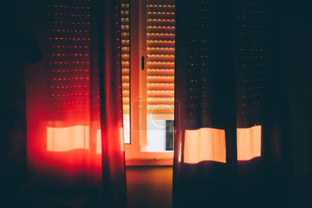 Foto de Por la tarde, el dormitorio está empapado en un cálido resplandor naranja a medida que la luz del sol se filtra a través de las persianas parcialmente dibujadas y se filtra a través de las cortinas rojas revoloteando en la brisa de la ventana abierta. - Imagen libre de derechos