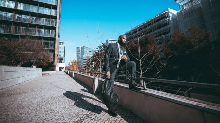 Photo for An elegant black man the outdoors relaxing, in the background, standing on the Portuguese cobblestone sidewalk wearing a full suit and carrying a weekender on a sunny and blue sky day - Royalty Free Image