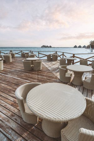 Photo for A vertical shot with selective focus on a garden table setting with cream-colored in rattan-effect style on a wooden floor and fencing overlooking the sea and the bungalows on a beautiful sunny day - Royalty Free Image