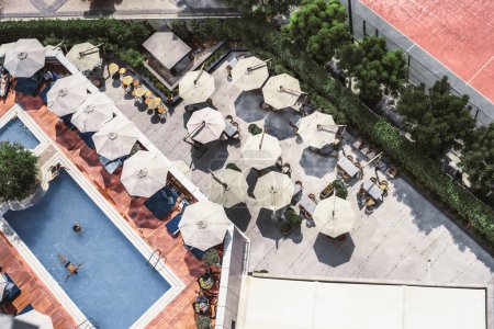 Foto de Disparo desde un dron, un bar al aire libre con múltiples sombrillas abiertas forma un paisaje agradable. Junto a la terraza se encuentra un exuberante jardín, mientras que el otro lado alberga una sala de estar con vistas a la piscina - Imagen libre de derechos