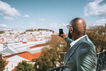 Téléchargez les photos : Capturez dans le focus sélectif un homme afro-touriste chauve et barbu dans une veste de costume vert pastel prend des photos de la ville d'un point de vue sur son smartphone par une journée lumineuse avec des nuages dans le ciel bleu - en image libre de droit