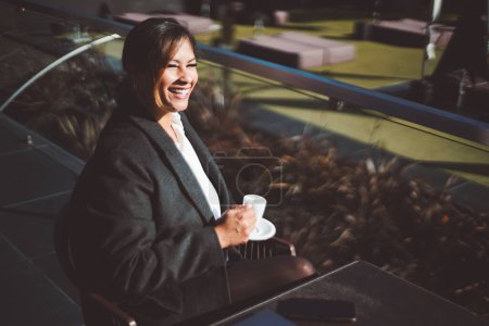 Téléchargez les photos : Assise sur une terrasse ensoleillée, une belle femme hispanique respire la joie et la confiance en prenant son café et en riant chaleureusement, revêtant un blazer noir chic. Son portable sur la table à côté d'elle - en image libre de droit