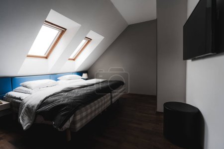 Foto de En gran angular, una acogedora cama con sábanas blancas y suaves cubiertas grises descansa contra una pared inclinada, bordeada por dos ventanas que permiten que la luz natural inunde el espacio. Frente a la cama, un televisor - Imagen libre de derechos