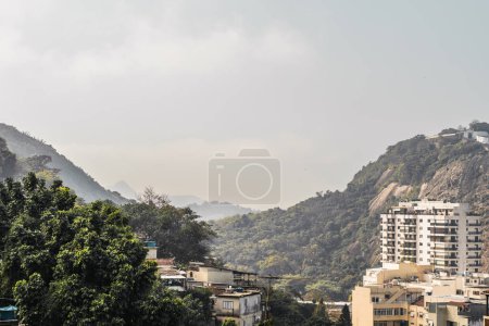 Téléchargez les photos : Vue depuis un appartement à Rio de Janeiro, entouré de montagnes luxuriantes couvertes d'arbres verts, d'humbles bâtiments parsèment le paysage ; La journée ensoleillée est accompagnée de nuages de brouillard léger - en image libre de droit