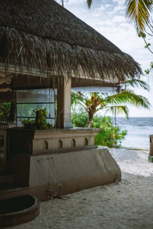 Téléchargez les photos : Journée ensoleillée dans un bar de plage de sable construit sur du grès avec un toit de chaume. Des cocotiers aux feuilles vertes peuvent être vus en arrière-plan, regardant la mer, apportant une touche naturelle à la scène - en image libre de droit