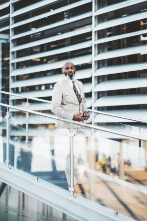 Photo for A vertical shot in Lisbon features a black man with a bald head and white beard, dressed in a stylish white suit and colorful striped tie. Stands by a seamless glass railing, exuding sophistication - Royalty Free Image